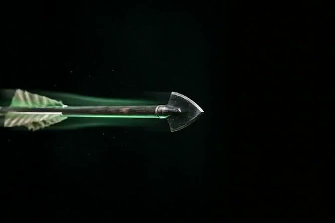 A broadhead arrow frozen mid-flight, trailing a green streak through the dark.